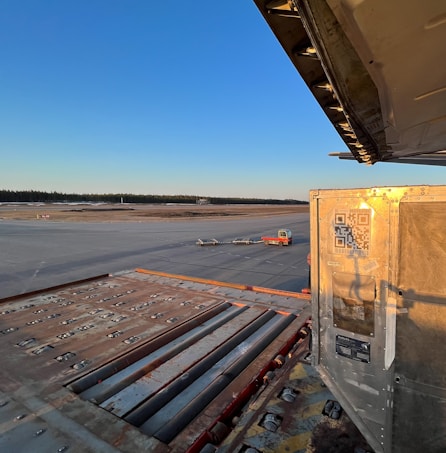A large open runway with a clear blue sky in the background. In the foreground, the edge of a cargo area is visible with metallic components and a QR code on a metal panel. A small red vehicle is parked on the tarmac along with a few trolley-like structures.