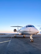 A sleek white private jet is parked on an airport runway under a clear blue sky. The aircraft's nose is prominently visible, highlighting its streamlined design. The background shows an expansive view of the runway, stretching out with no obstructions.