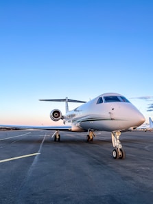 A sleek white private jet is parked on an airport runway under a clear blue sky. The aircraft's nose is prominently visible, highlighting its streamlined design. The background shows an expansive view of the runway, stretching out with no obstructions.