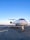 A sleek white private jet is parked on an airport runway under a clear blue sky. The aircraft's nose is prominently visible, highlighting its streamlined design. The background shows an expansive view of the runway, stretching out with no obstructions.