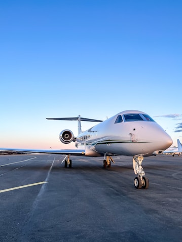 A sleek white private jet is parked on an airport runway under a clear blue sky. The aircraft's nose is prominently visible, highlighting its streamlined design. The background shows an expansive view of the runway, stretching out with no obstructions.