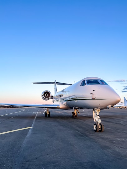A sleek white private jet is parked on an airport runway under a clear blue sky. The aircraft's nose is prominently visible, highlighting its streamlined design. The background shows an expansive view of the runway, stretching out with no obstructions.