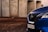 The image features a close-up view of the front part of a blue Nissan vehicle, with focus on the grille and headlight. The background includes concrete structures and a dirt ground, suggesting an industrial setting.