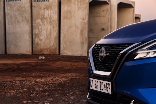 The image features a close-up view of the front part of a blue Nissan vehicle, with focus on the grille and headlight. The background includes concrete structures and a dirt ground, suggesting an industrial setting.