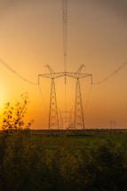 Tall metal transmission towers stretch into the distance, carrying electrical lines across a flat landscape. The scene is set against a warm, golden sunset sky with a soft gradient from orange to yellow. The foreground features dense foliage and green fields, adding a natural contrast to the industrial structures.