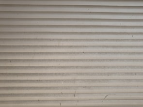 A series of horizontal metal slats form a shutter or garage door, featuring a light gray color with various small marks and stains across its surface.