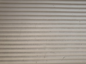 A series of horizontal metal slats form a shutter or garage door, featuring a light gray color with various small marks and stains across its surface.