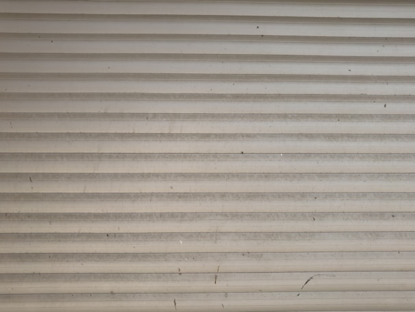 A series of horizontal metal slats form a shutter or garage door, featuring a light gray color with various small marks and stains across its surface.
