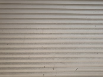 A series of horizontal metal slats form a shutter or garage door, featuring a light gray color with various small marks and stains across its surface.