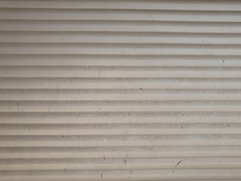 A series of horizontal metal slats form a shutter or garage door, featuring a light gray color with various small marks and stains across its surface.