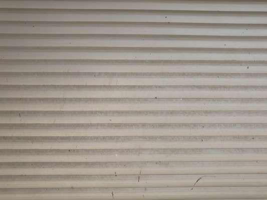 A series of horizontal metal slats form a shutter or garage door, featuring a light gray color with various small marks and stains across its surface.