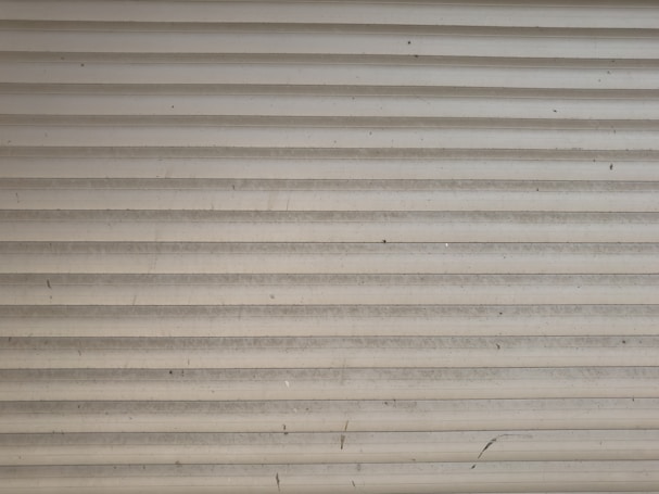 A series of horizontal metal slats form a shutter or garage door, featuring a light gray color with various small marks and stains across its surface.