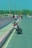 A motorcycle is parked on the side of a road next to a walkway with people walking. The road is wide and curves gently into the distance, lined with tall lampposts and bordered by a barrier painted with red and white stripes. Several people are visible on the sidewalk, and there are vehicles, including a three-wheeler, on the road.