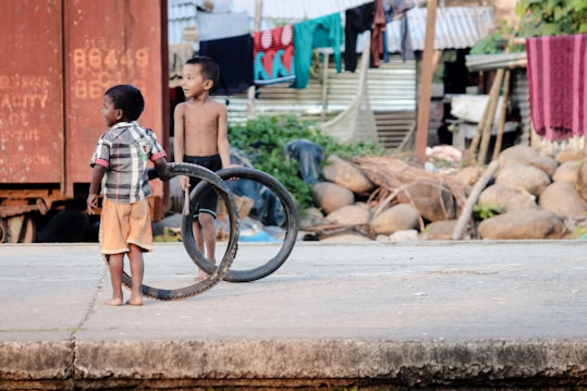 Two young boys are outdoors in a rural setting. One boy is shirtless and wearing shorts, while the other is dressed in a checkered shirt and orange shorts. They are holding bicycle tires as playthings. In the background, there are stacked rocks, metal sheets, and clothes hanging on a line, suggesting a residential area.