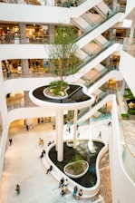 A modern shopping mall interior with multiple floors connected by escalators. The central feature is a large, elevated indoor garden with a tree, surrounded by water and seating areas. Shoppers and visitors can be seen walking around the open, bright space, highlighting the mall's airy design.
