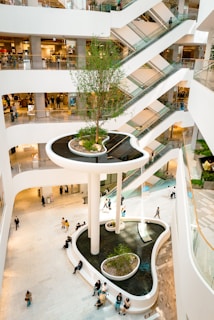 A modern shopping mall interior with multiple floors connected by escalators. The central feature is a large, elevated indoor garden with a tree, surrounded by water and seating areas. Shoppers and visitors can be seen walking around the open, bright space, highlighting the mall's airy design.