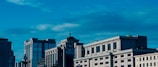 Several modern office buildings with large glass windows stand under a bright blue sky with scattered clouds. The architecture features a mix of contemporary and classical design elements, highlighted by geometric patterns and rectilinear forms.