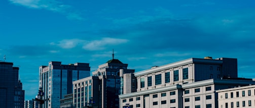 Several modern office buildings with large glass windows stand under a bright blue sky with scattered clouds. The architecture features a mix of contemporary and classical design elements, highlighted by geometric patterns and rectilinear forms.