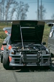 A modified sports car with its hood open, revealing its engine. The car features bold graphics in orange and grey and appears to be set in an outdoor environment, with blurred trees in the background.