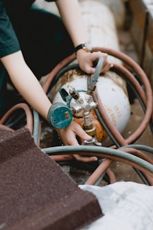 A person is adjusting a nozzle or valve on a gas cylinder using both hands. The scene includes a complex arrangement of hoses encircling the cylinder. The individual wears a dark shirt and a wristwatch. The setting appears to be industrial or utilitarian, with various equipment partially visible.