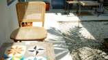 A wooden chair with a woven backrest is positioned next to a small table featuring colorful geometric tiles. The area is well-lit, casting shadows across a decorative pebble garden, with several chairs and tables visible in the background.