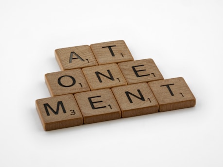 Wooden Scrabble tiles arranged to spell the word 'ATONEMENT', each with a number indicating their point value.