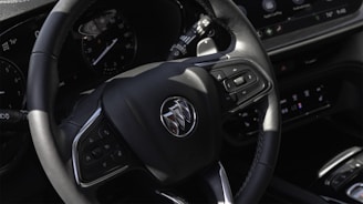A close-up view of a car's steering wheel featuring a brand emblem. The wheel has multiple control buttons and is surrounded by a dashboard with instrument gauges and a digital display.