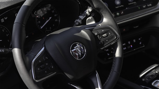 A close-up view of a car's steering wheel featuring a brand emblem. The wheel has multiple control buttons and is surrounded by a dashboard with instrument gauges and a digital display.