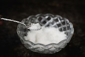 A clear, intricately designed glass bowl holds a quantity of white crystalline salt. A metallic spoon is poised above the bowl, containing a small amount of the salt.