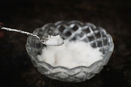A clear, intricately designed glass bowl holds a quantity of white crystalline salt. A metallic spoon is poised above the bowl, containing a small amount of the salt.