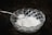 A clear, intricately designed glass bowl holds a quantity of white crystalline salt. A metallic spoon is poised above the bowl, containing a small amount of the salt.