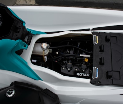 A close-up view of a mechanical engine inside a watercraft, featuring branding that reads 'ROTAX'. The engine is housed within a white and blue-green compartment, showing various components such as hoses and caps. The color scheme is primarily metallic with hints of plastic casings.