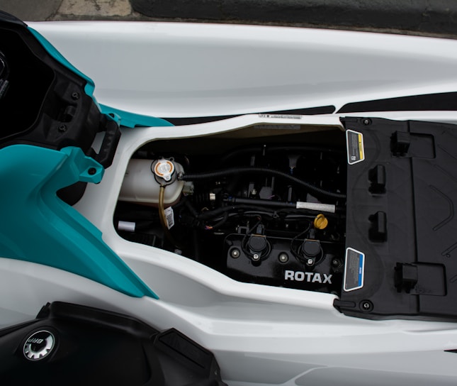A close-up view of a mechanical engine inside a watercraft, featuring branding that reads 'ROTAX'. The engine is housed within a white and blue-green compartment, showing various components such as hoses and caps. The color scheme is primarily metallic with hints of plastic casings.