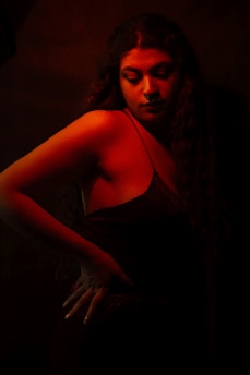 A woman with long hair is posed against a dark background, illuminated dramatically by red and orange lighting. Her eyes are softly closed and her arm is positioned on her hip, adding to a sense of confidence and contemplation.