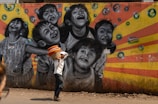 A vibrant street mural depicting joyful children laughing and playing, with bubbles surrounding them. The background features a burst of orange and yellow stripes. In the foreground, a man is walking by, carrying a basket filled with colorful cloth items on his head. The scene combines art and daily life in an urban setting.