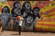 A vibrant street mural depicting joyful children laughing and playing, with bubbles surrounding them. The background features a burst of orange and yellow stripes. In the foreground, a man is walking by, carrying a basket filled with colorful cloth items on his head. The scene combines art and daily life in an urban setting.