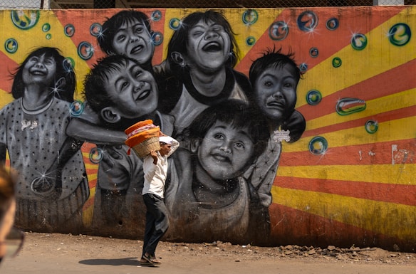A vibrant street mural depicting joyful children laughing and playing, with bubbles surrounding them. The background features a burst of orange and yellow stripes. In the foreground, a man is walking by, carrying a basket filled with colorful cloth items on his head. The scene combines art and daily life in an urban setting.