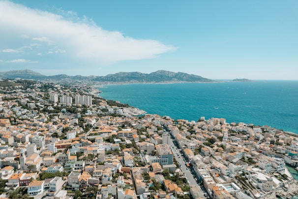 A coastal cityscape with numerous buildings clustered together, overlooking a vibrant blue sea. The horizon shows a range of mountains under a clear blue sky. Tall apartment complexes are visible near the shore, interspersed with smaller residential structures.