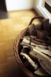 A wicker basket filled with neatly stacked pieces of firewood against a wooden floor background. The basket is woven and has a handle, positioned near a light source that creates soft shadows.
