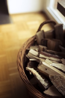 A wicker basket filled with neatly stacked pieces of firewood against a wooden floor background. The basket is woven and has a handle, positioned near a light source that creates soft shadows.