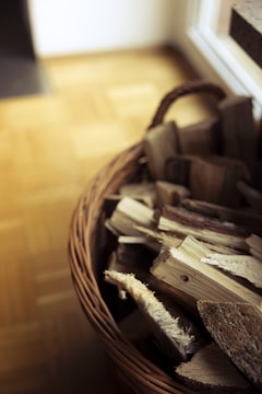 A wicker basket filled with neatly stacked pieces of firewood against a wooden floor background. The basket is woven and has a handle, positioned near a light source that creates soft shadows.