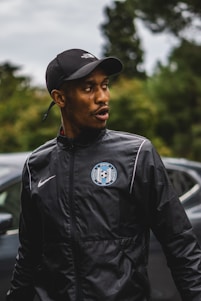 A person wearing a black Nike jacket with a sports emblem, along with a black baseball cap, stands outdoors. The background includes a blurred view of trees and a car.