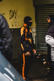 A person wearing a black and orange tracksuit and a balaclava stands facing another individual in a dark clothing and hat. They appear to be in an urban environment, possibly an alley, with graffiti on the wall and litter on the ground.