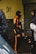 A person wearing a black and orange tracksuit and a balaclava stands facing another individual in a dark clothing and hat. They appear to be in an urban environment, possibly an alley, with graffiti on the wall and litter on the ground.