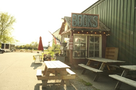 A small rustic bookshop with a sign that reads 'BOOKS' situated along the side of a green metal building. Several wooden picnic tables are positioned in front of the shop. An open umbrella stands beside a small table decorated with some items near the road. The scene is sunlit and appears quiet and inviting, with a few potted plants adding to the charm.