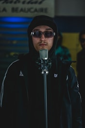 A person wearing dark sunglasses and a hooded sweatshirt stands in front of a microphone. The background has a blurred sign and appears to be dimly lit with a blue and yellow color scheme.