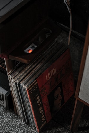 A collection of vinyl records stored upright with a prominent album cover featuring the word 'LATIN' and an illustration of a woman's face. The scene is dimly lit, and there's an electronic device and a power strip nearby.