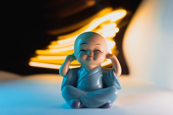 A small figurine of a smiling monk sitting in a meditative pose with hands covering its ears. The background features abstract streaks of yellow light against a dark setting, creating a striking contrast.