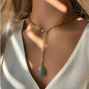 A close-up shot of a woman's neck wearing a gold chain necklace with an elongated oval green gemstone pendant. The background is softly lit, casting delicate shadows and emphasizing the elegance of the jewelry.