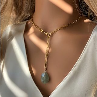 A close-up shot of a woman's neck wearing a gold chain necklace with an elongated oval green gemstone pendant. The background is softly lit, casting delicate shadows and emphasizing the elegance of the jewelry.
